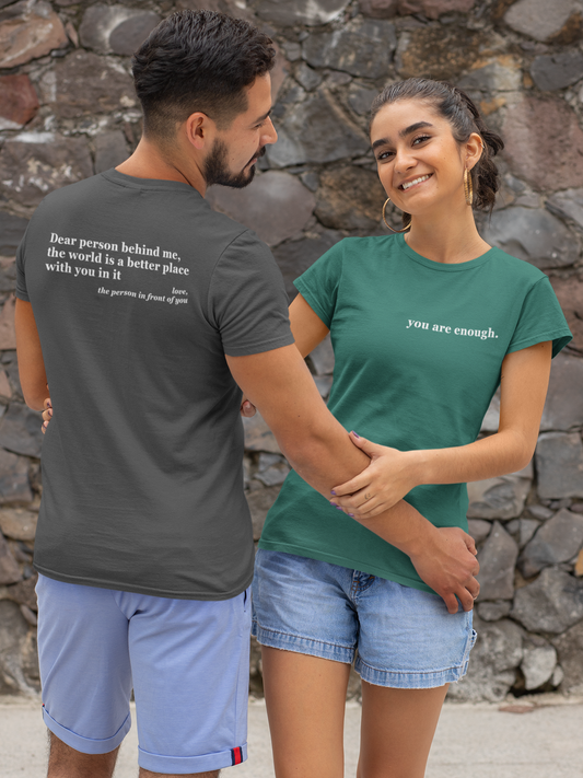 Two people standing close together, one wearing a gray t-shirt with a motivational quote and the other wearing a green t-shirt with 'you are enough' text, against a stone wall background.